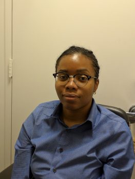 Ashley is a black female with dark brown eyes, braided hair, and oval-shaped glasses. Wearing a blue long sleeve collared shirt.