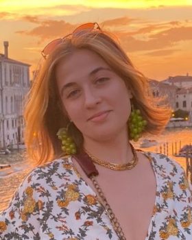 Picture of Ann DeAngelis, a white female with blue eyes, a light brown hair with a short bob haircut. Wearing a white dress with a green, yellow and orange flower pattern, a gold necklace and green grape earrings. Ann is pictured in front of a sunset background.