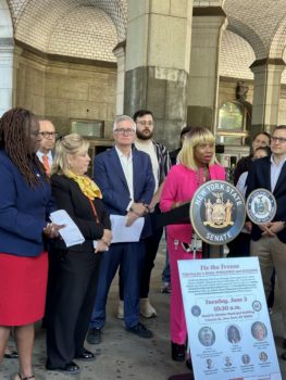Dr. Sharon McLennon Wier in pink speaking at an outdoor podium surrounded by elected officials and advocates.