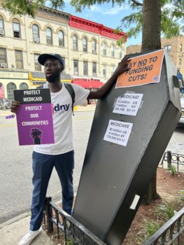 CIDNY's Community Organizer Mbacke stands next to a symbolic casket at a Medicaid rally in Brooklyn.
