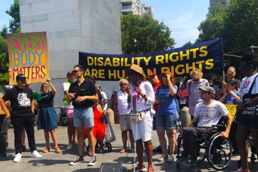 A group of people are gathered outdoors with CIDNY's Dr. Sharon McLennon Wier speaking in front of a banner that reads "Disability Rights Are Human Rights."